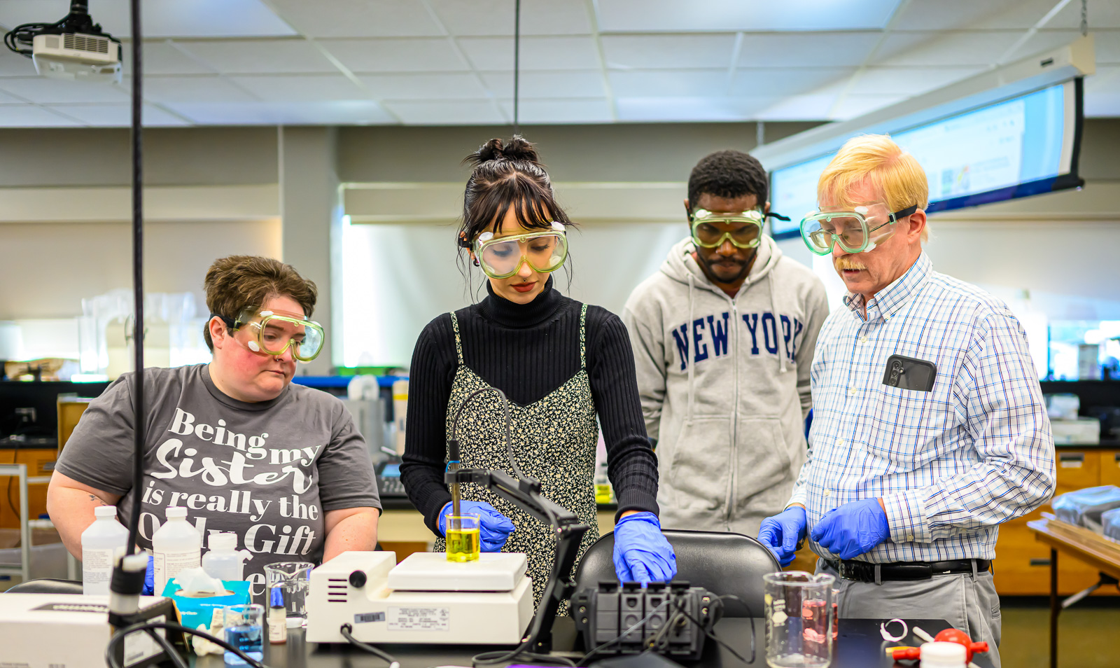 Three Bristol students working with their professor, all wearing goggles and gloves. One student is handling lab equipment while the other two watch.