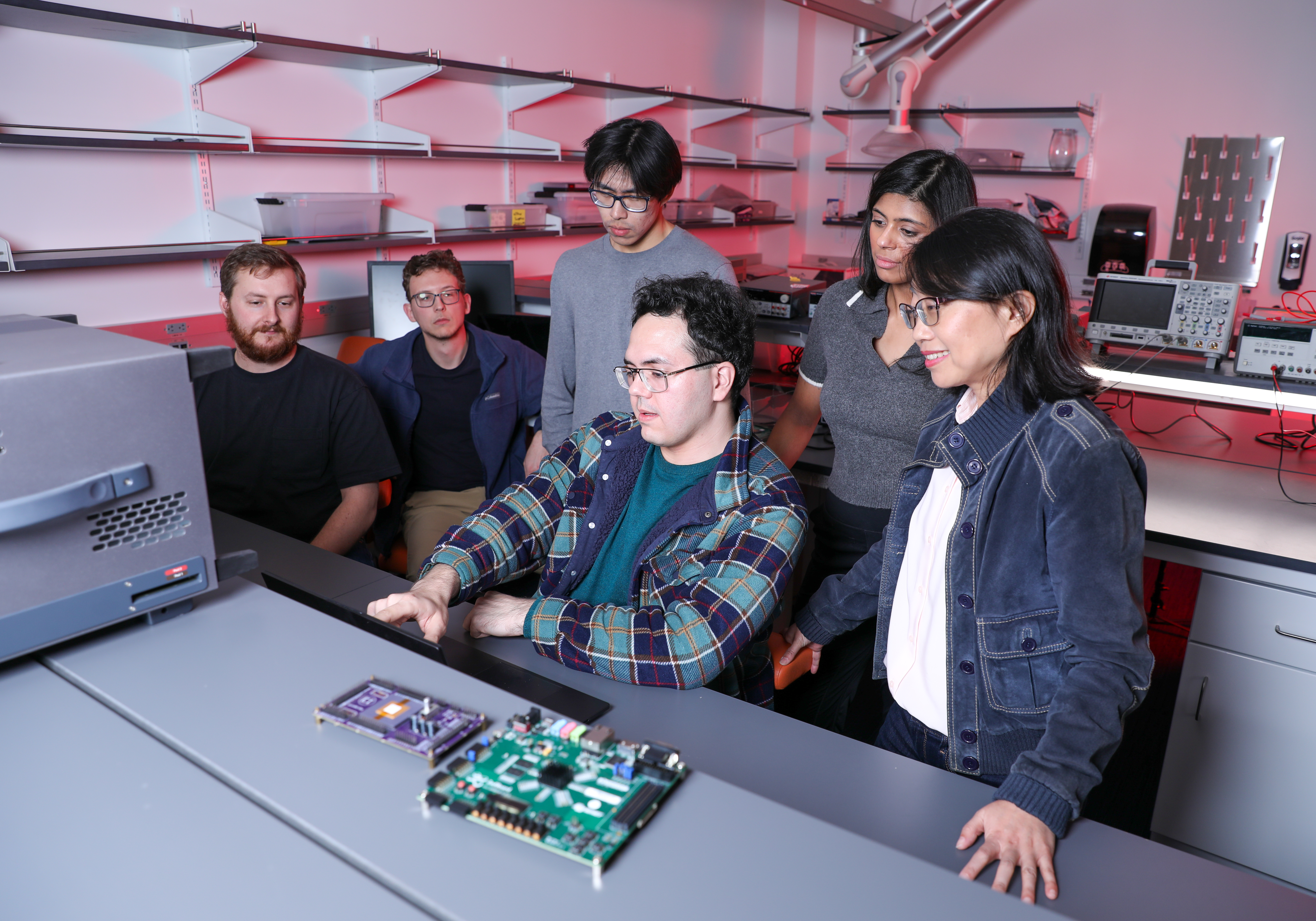 Dr. Helen "Hai" Li stands with a group of 5 students in her lab around a computer in the Wilkinson building on campus, as one student dictates to the group about the current state of his research. There is a motherboard in the foreground of the image.