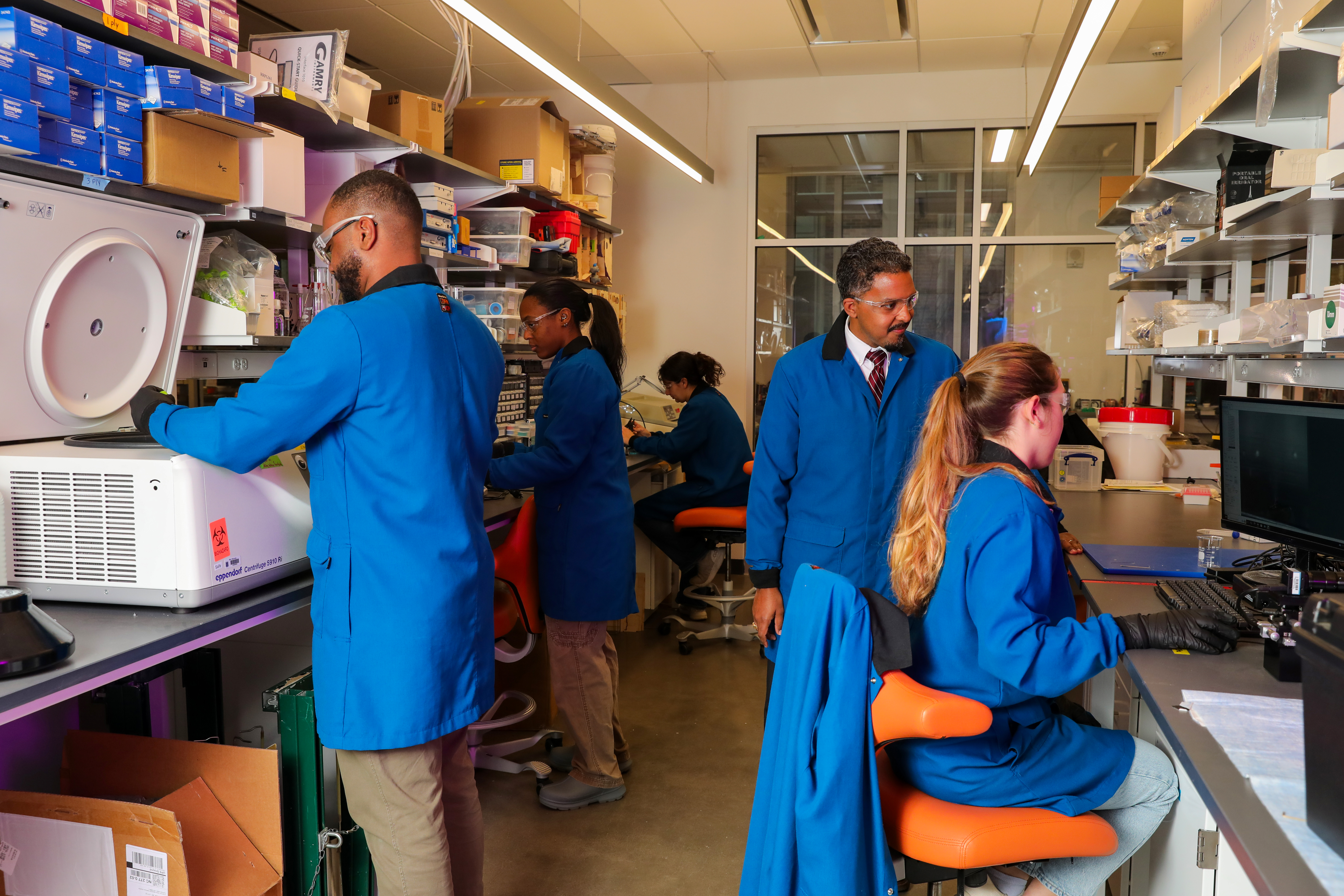Four students stand at different workstations in a lab while Professor Andrew D Jones speaks to a student at a computer. The image takes place on the second floor of the Wilkinson building and was taken January 6, 2025.