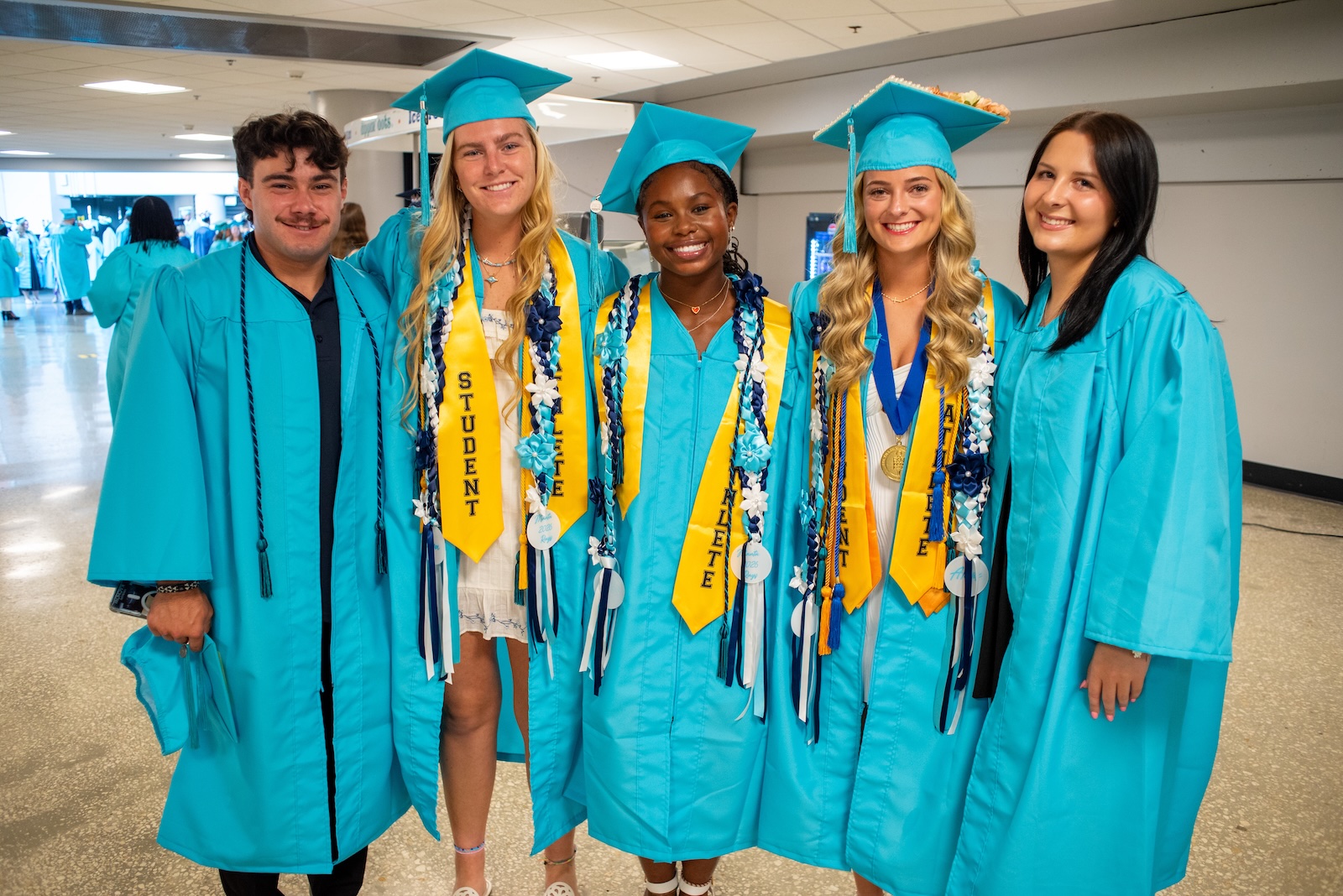 Five young adults pose together at an FSCJ graduation ceremony, with all wearing teal gowns and some wearing teal caps and various honor cords.