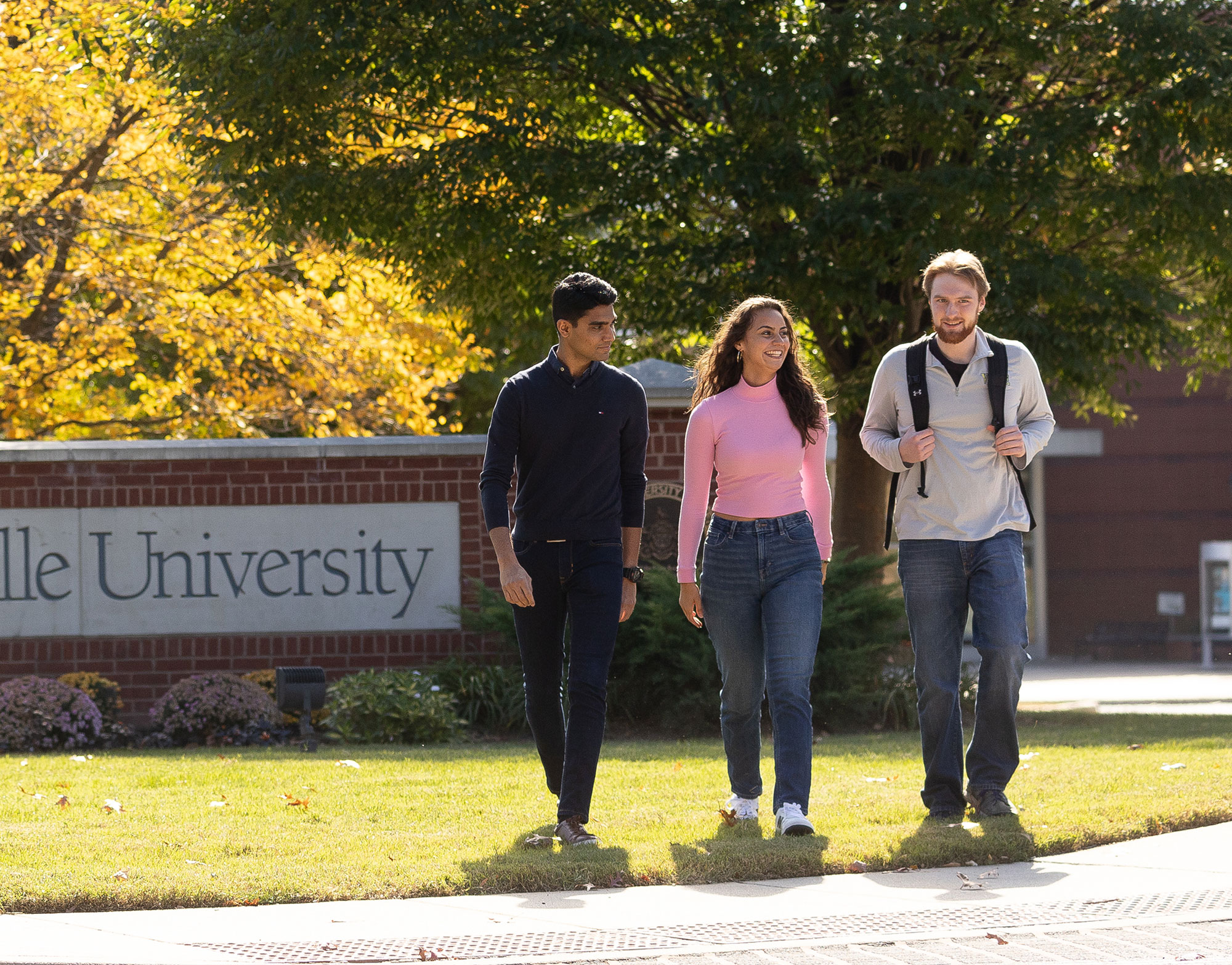 three students walking on campus with a brick Millersville University sign in the background.