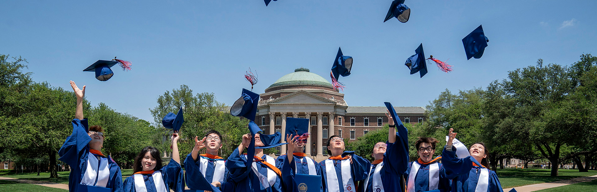 Recent SMU Grads tossing caps in air in front of Dallas Hall.