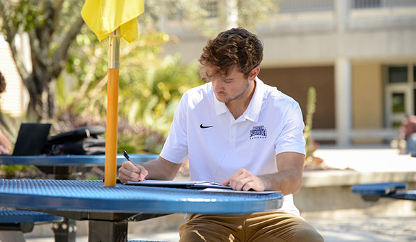Male student sitting at outside table writing on notepad