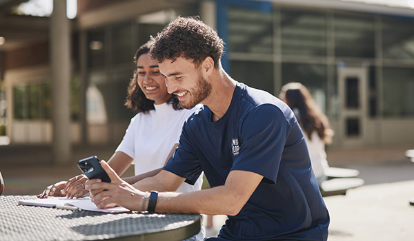 Male and female students at outside table smiling and looking at cell phone