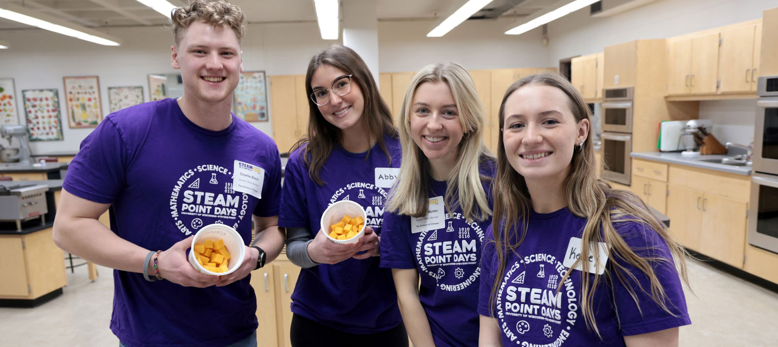 Group of four students standing in a lab with matchin purple shirts with two students holding food in cups. 